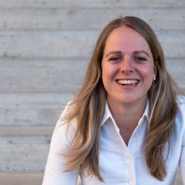 A woman with long blonde hair wearing a white shirt is smiling while sitting in front of a concrete staircase.