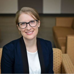 A person wearing glasses and a blazer sits on a patterned sofa, smiling at the camera in a lounge area with more sofas in the background.