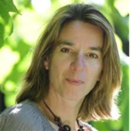 A woman with shoulder-length light brown hair is outdoors, wearing a light sweater and a necklace, with green foliage in the blurred background.