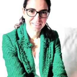 Woman with dark hair and glasses wearing a textured green blazer sits and smiles at the camera, with a bright background and a partial framed picture visible above her.