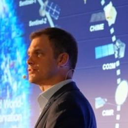 Man wearing a suit and headset microphone speaks in front of a screen displaying satellite images and labels related to Earth observation.