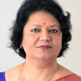 A woman with short, curly black hair, wearing earrings, a gold chain, a bindi on her forehead, and a saree with a pink border, poses against a plain light background.