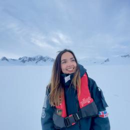 A person in a dark jacket and red life vest stands smiling in a snowy, mountainous landscape under a cloudy sky.