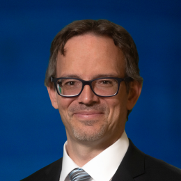 A man with short brown hair and glasses, wearing a dark suit, white shirt, and striped tie, smiles in front of a blue background.