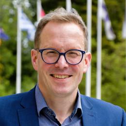 A man wearing blue glasses and a blue blazer smiles outdoors, with several out-of-focus flags and greenery in the background.