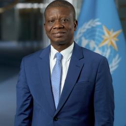 A man in a blue suit and tie stands in front of a blue flag with a gold star and white emblem, indoors.