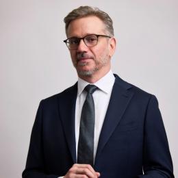 Man wearing glasses and a dark suit with a white shirt and tie stands against a plain light background, looking at the camera with hands clasped.