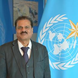 Man in a dark grey suit and patterned tie standing in front of a blue World Meteorological Organization flag.
