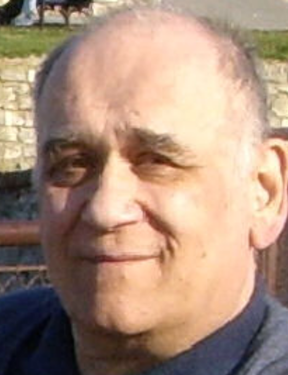 A man in a blue shirt is smiling in front of a bridge.