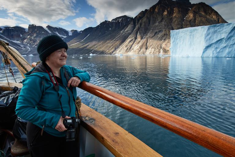 A person in a blue jacket and black beanie holds a camera and stands on a boat, looking at a large iceberg and mountainous landscape in the background.