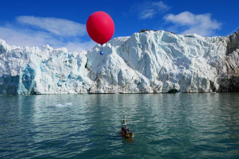 A large red balloon hovers above a small boat near an iceberg in a calm sea under a clear sky.