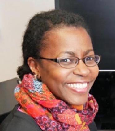 A woman wearing glasses and a scarf smiles in front of a computer.