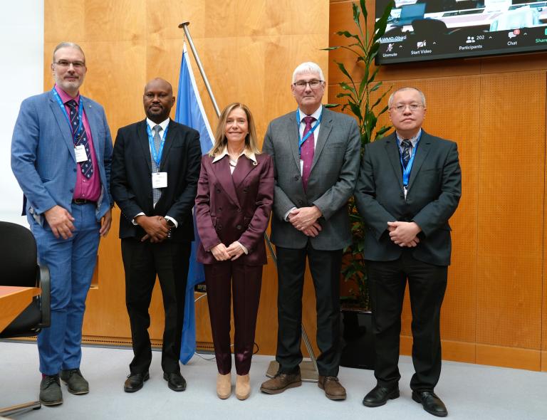 Five professionals standing in a conference room with a un flag in the background, smiling at the camera.