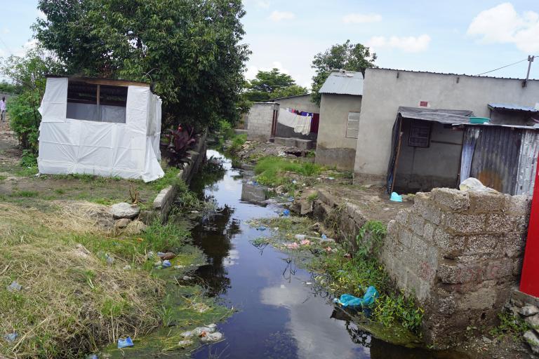 A stream of water running through an informal settlement with litter scattered around and simple structures on either side.
