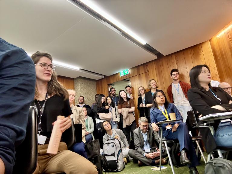 A diverse group of people, some seated and some standing, attentively listens to a presentation in a modern, well-lit conference room with wood-paneled walls.