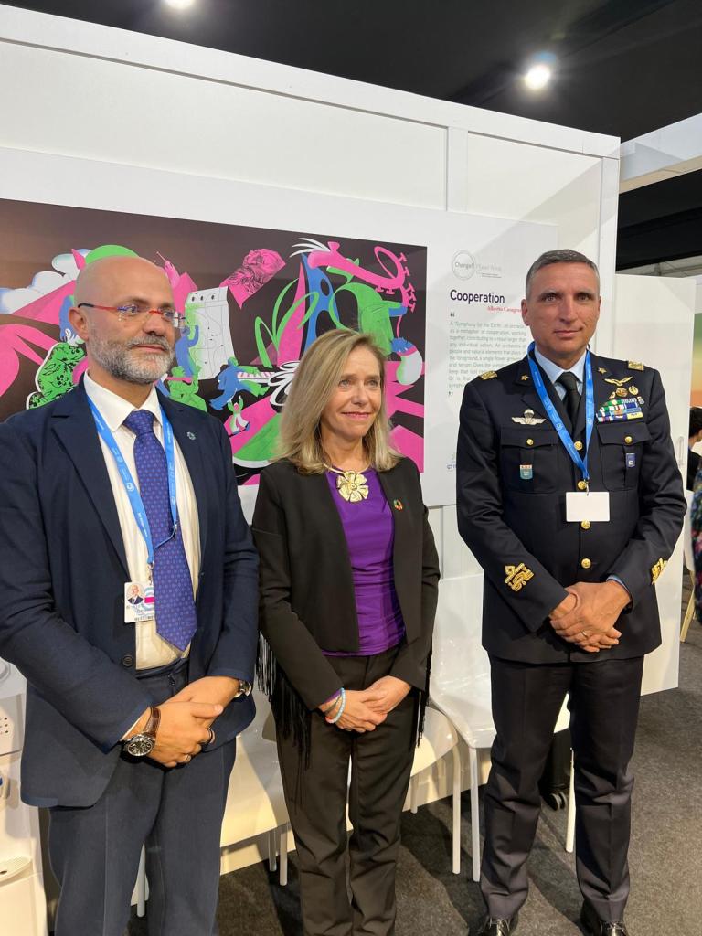 Three people stand side by side in formal attire at an event, with a colorful display and text about cooperation in the background.