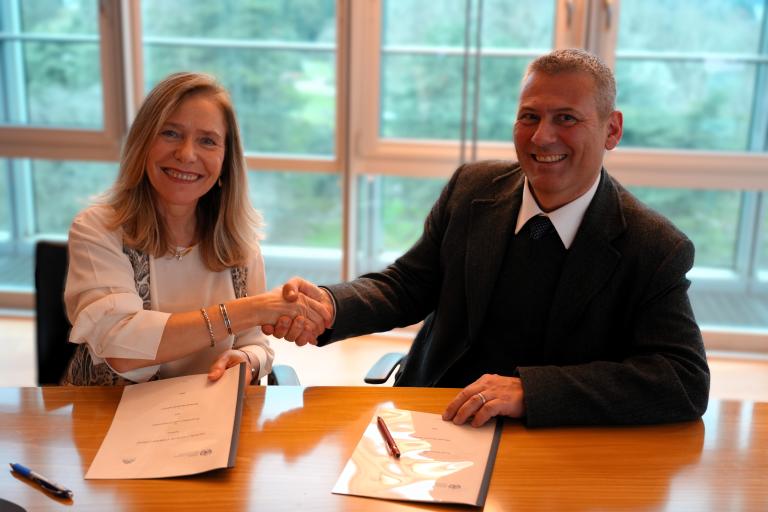 Two people are sitting at a table, shaking hands over signed documents. There are pens placed on the papers. The background shows large windows with a view of greenery.
