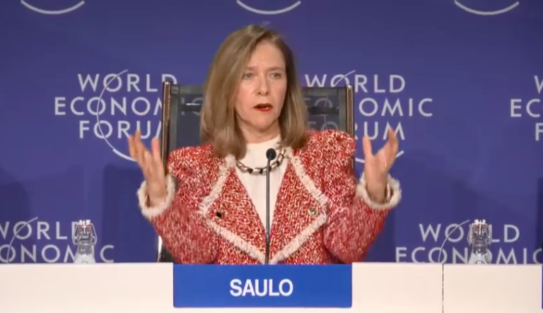A person gestures while speaking at a World Economic Forum panel, seated in front of a backdrop with the event's logo. The nameplate reads "SAULO.