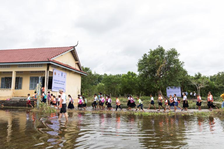 Children in school uniforms walk in a line through floodwaters next to a building with a red roof. Trees and a cloudy sky are in the background.