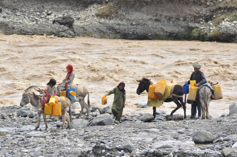People and donkeys carrying yellow containers navigate a rocky riverbank with a fast-flowing river in the background.
