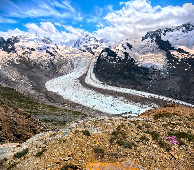 Mountainous landscape with a winding glacier flowing through rocky terrain. Snow-capped peaks are visible under a partly cloudy sky. Sparse vegetation is present in the foreground.