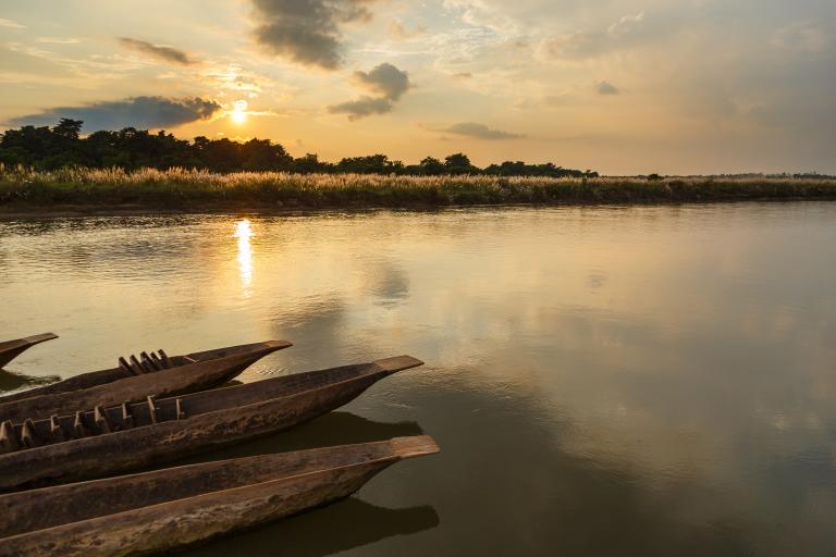 Canoes are docked by a calm river under a cloudy sky at sunset, with grass and trees in the background.