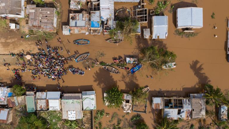Aerial view of a flooded village with submerged buildings and boats. A large group of people gathers on dry ground, surrounded by muddy water. Trees and damaged structures are visible.