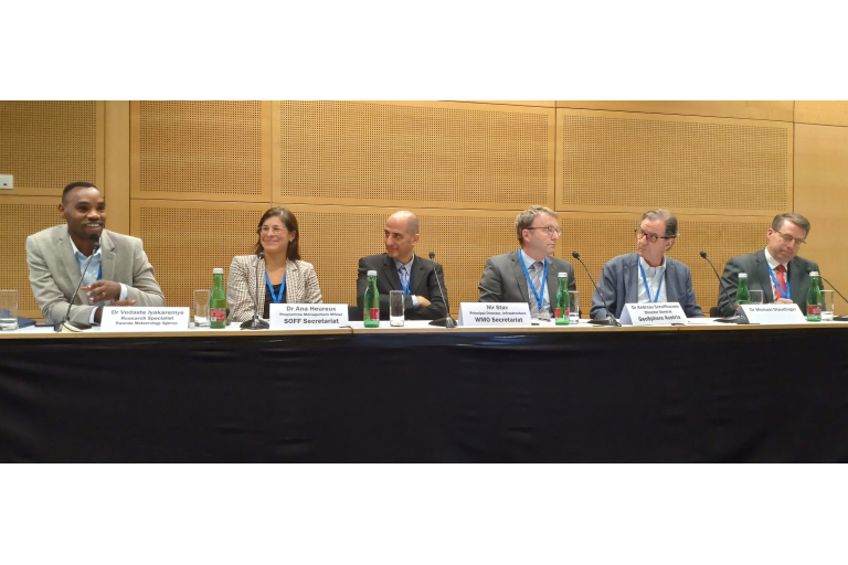 Six people sit behind a panel table with microphones, speaking at a conference. They have name tags and water bottles in front of them.