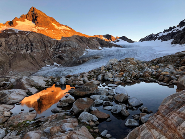 Mountain with a snow-covered slope and glacier reflecting in a small pond surrounded by rocks at sunset.