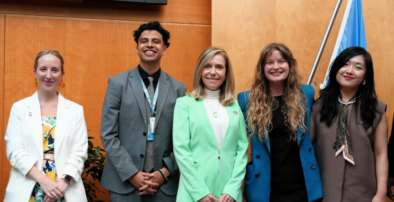 Five people are standing side by side in front of a wooden paneled wall and a flag, smiling for a group photo.