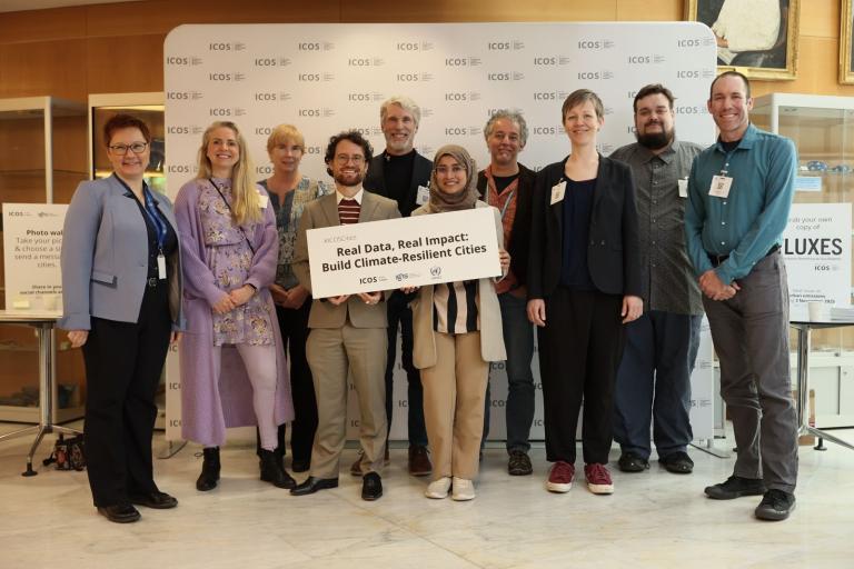 A diverse group of people stands together, smiling, holding a sign reading, "Real Data, Real Impact: Build Climate-Resilient Cities," at an event with a branded backdrop.