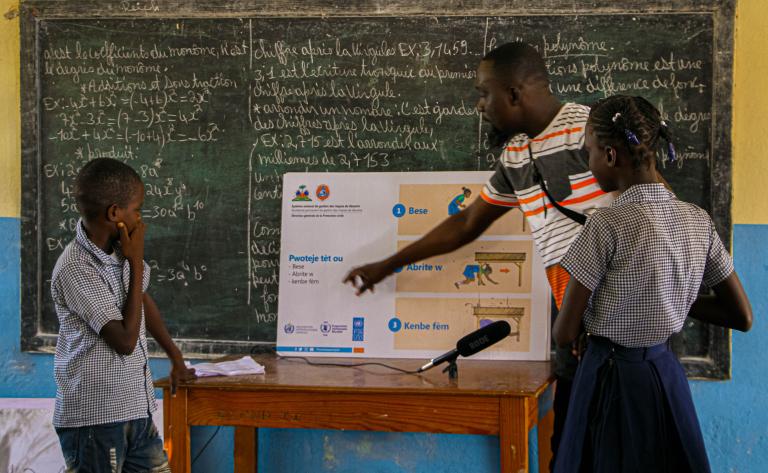 Teacher explaining a safety poster to two students in front of a chalkboard in a classroom. The chalkboard is filled with text, and a microphone is visible on the table.