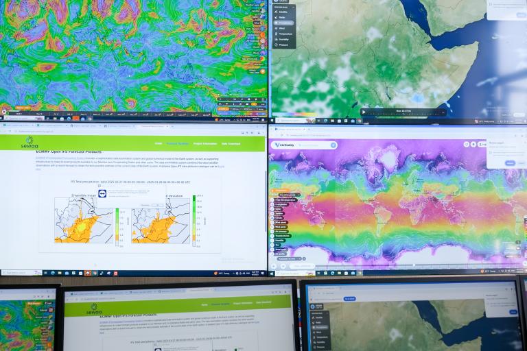 Six computer monitors display various weather, wind, and satellite maps, including charts and global patterns, for different regions and data analysis.