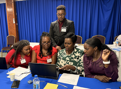 Four women gather around a table, focused on a laptop. A man, focused on the laptop, stands behind them. Papers and bottles are scattered on the table. A blue curtain and chairs are in the background.