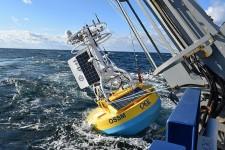 A buoy equipped with solar panels and instruments is being deployed from a ship into the ocean on a bright, partly cloudy day.