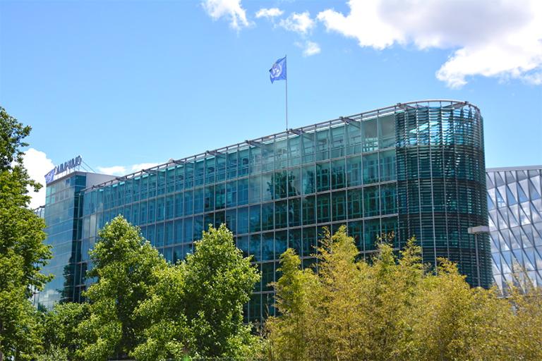A modern glass office building with a blue company flag on the roof, partially obscured by trees under a bright sky with clouds.