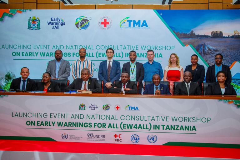 A group of people, some seated and some standing, at a workshop event. A banner in the background reads "Launching Event and National Consultative Workshop on Early Warnings for All in Tanzania.