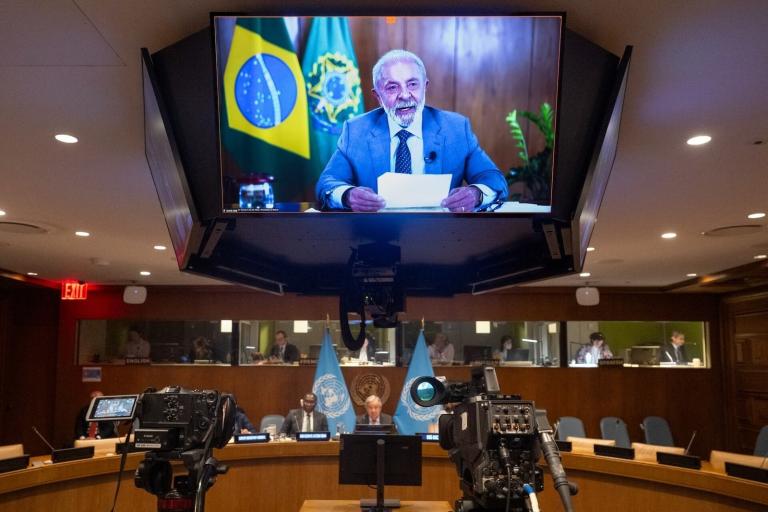 A large screen shows a man speaking during a video conference at a UN meeting; Brazilian and UN flags are visible, with cameras recording in the foreground.