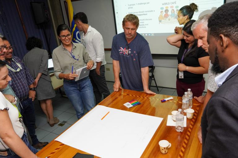 A group of people stands around a table with a large sheet of paper, discussing in a meeting room with a presentation projected on a screen in the background.