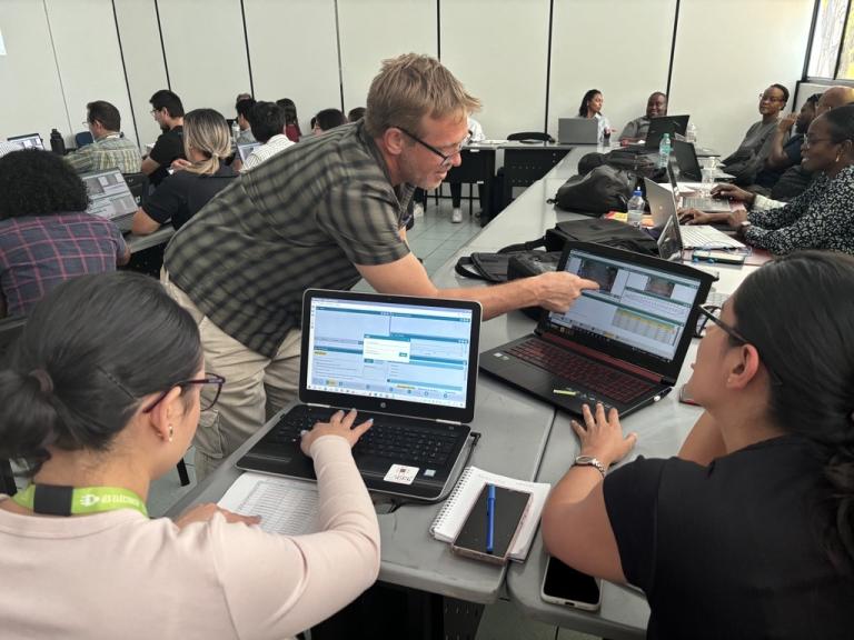 A man leans over to assist two women working on laptops in a classroom setting; several others are seated at tables working on computers in the background.