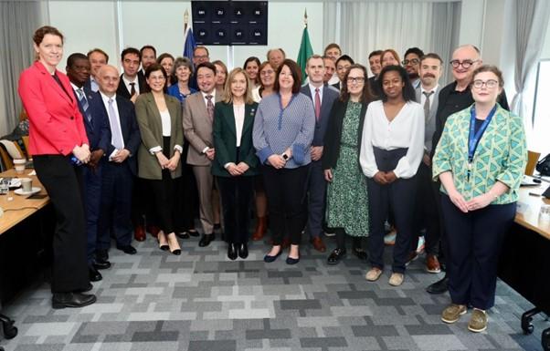 A group of around 25 people poses for a photo in a conference room, standing in front of desks and flags, dressed in business attire.