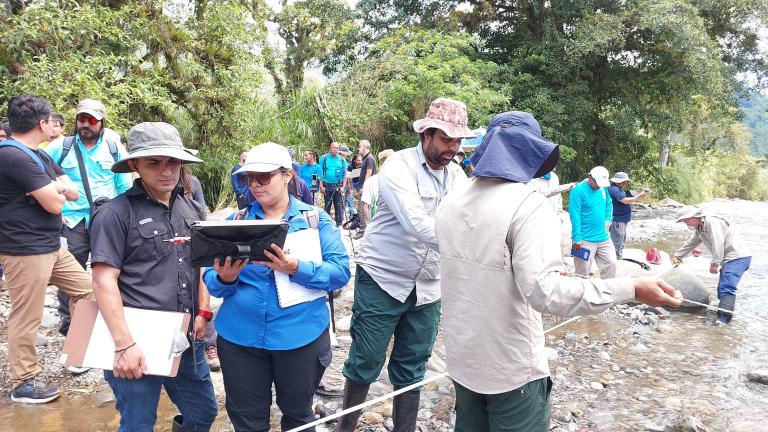 A group of people, some with clipboards and equipment, conduct fieldwork and take measurements by a rocky river in a forested area.