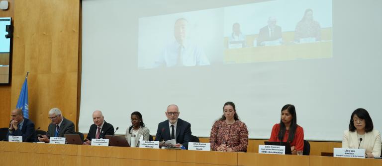 Seven people sit at a conference table with microphones, while three others appear on a screen behind them, participating virtually. A blue flag is visible to the left.
