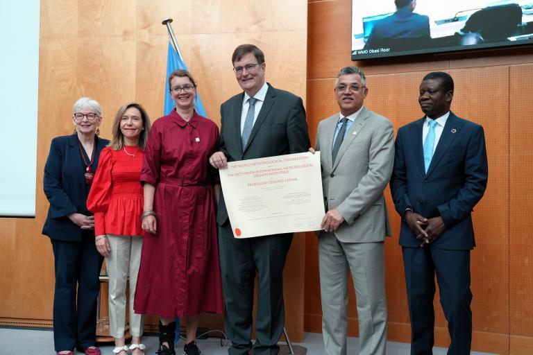 Six people stand indoors, with one person in the center holding a large certificate. A UN flag is in the background. They are formally dressed and posing for a group photo.
