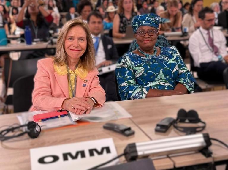 Two women sit at a conference table with documents and microphones, surrounded by other attendees. A sign marked "OMM" is visible in the foreground.