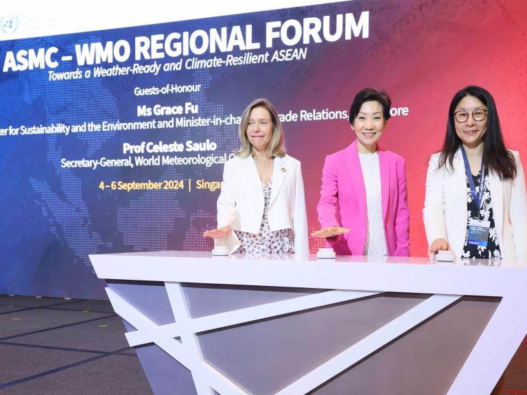 Three women stand behind a podium at the ASMC-WMO Regional Forum in Singapore, with a large event banner displayed in the background.