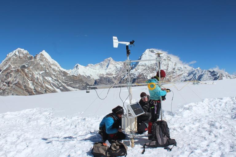 Three people set up scientific equipment on a snowy mountain with high peaks in the background under a clear blue sky.