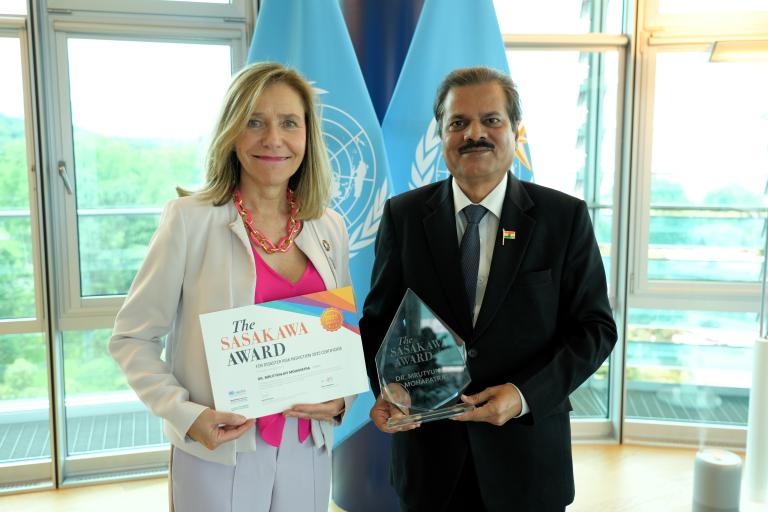A woman holds a certificate and a man holds a glass award; they stand in front of UN flags and large windows.