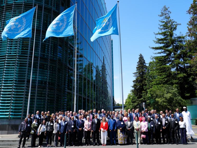 A large group of people pose for a photo outside a glass building with three United Nations flags, surrounded by trees under a clear sky.