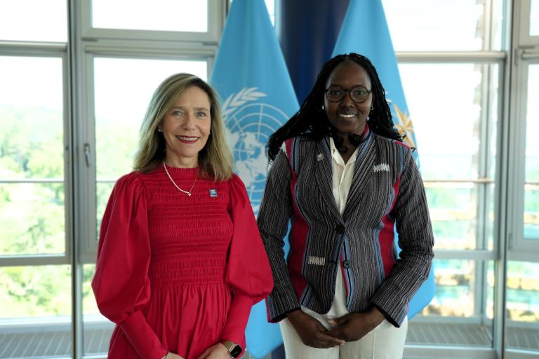 Two women stand side by side indoors in front of large windows and two blue United Nations flags. One woman wears a red dress, and the other wears a striped jacket and white shirt.